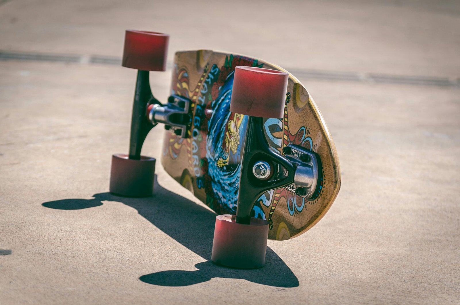 Close-up shot of a colorful longboard with red wheels on the pavement, casting a shadow.